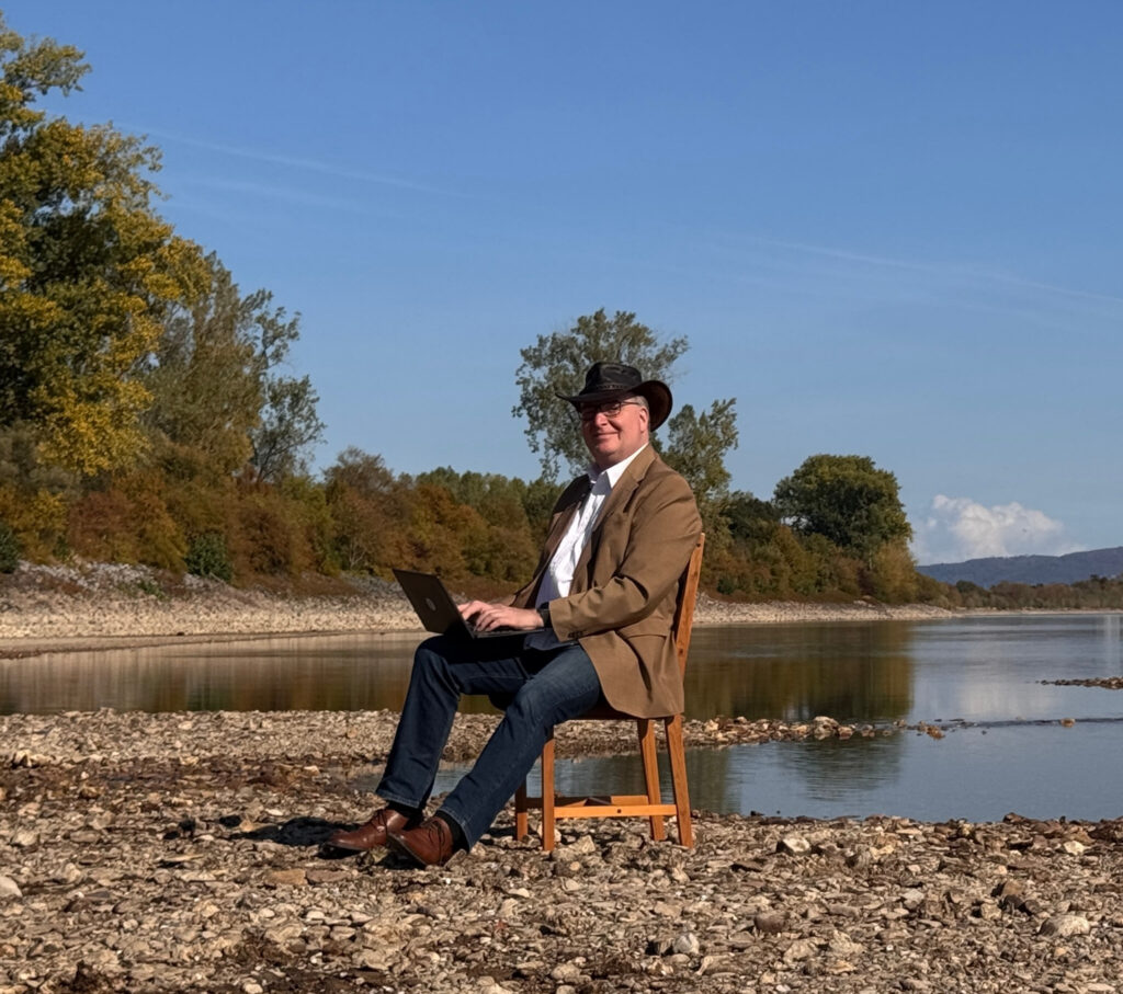Clark Ahten auf einem Stuhl am steinigen Rheinufer sitzend, direkt am Wasser, Laptop auf dem Schoss, Hut auf.